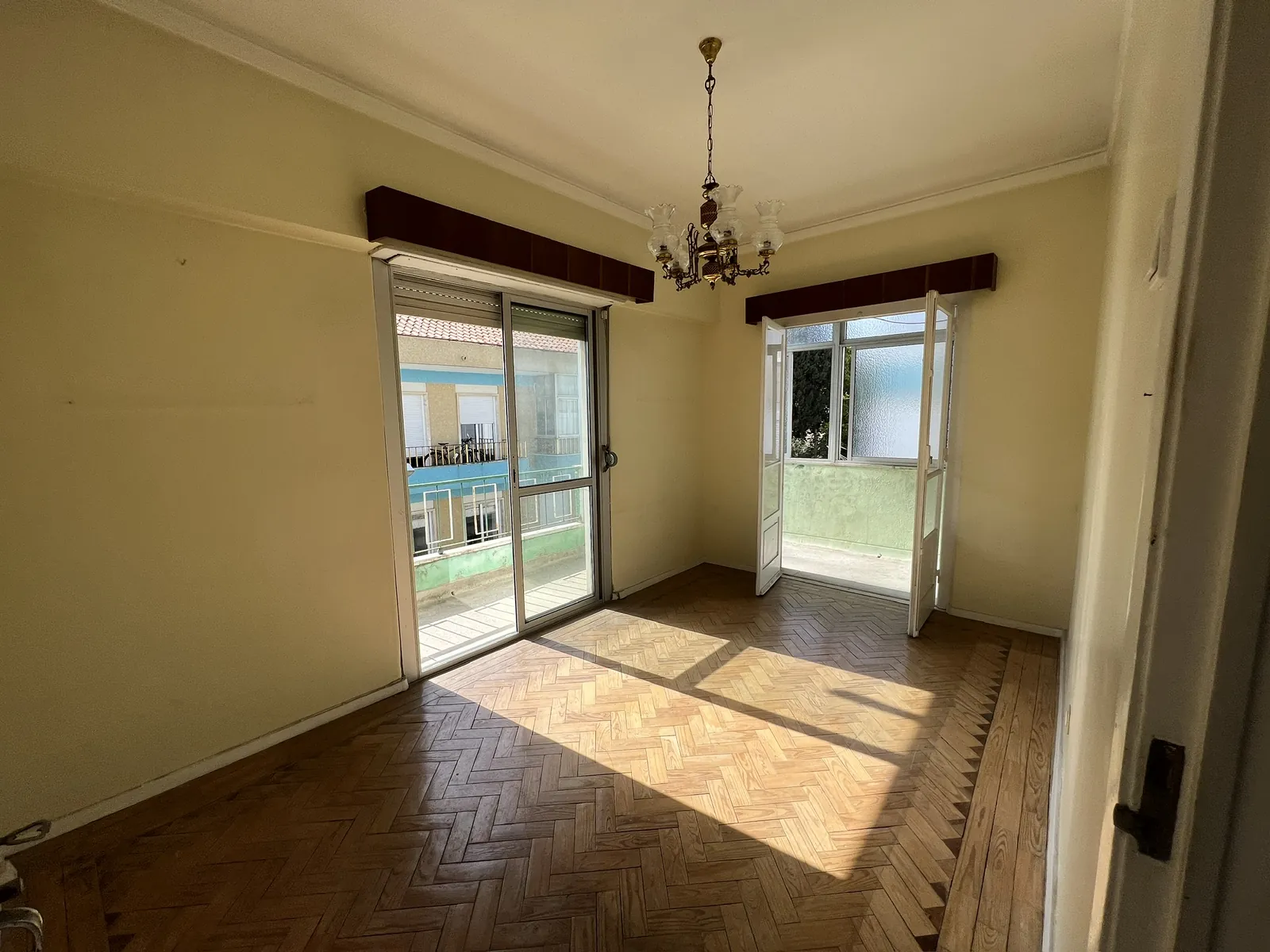 Sala antes da obra, vazia, com pavimento em espinha e candeeiro antigo